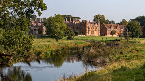A view of the house at Charlecote Park from across the River Avon on a sunny day, with greenery on the banks and trees in front of the house
